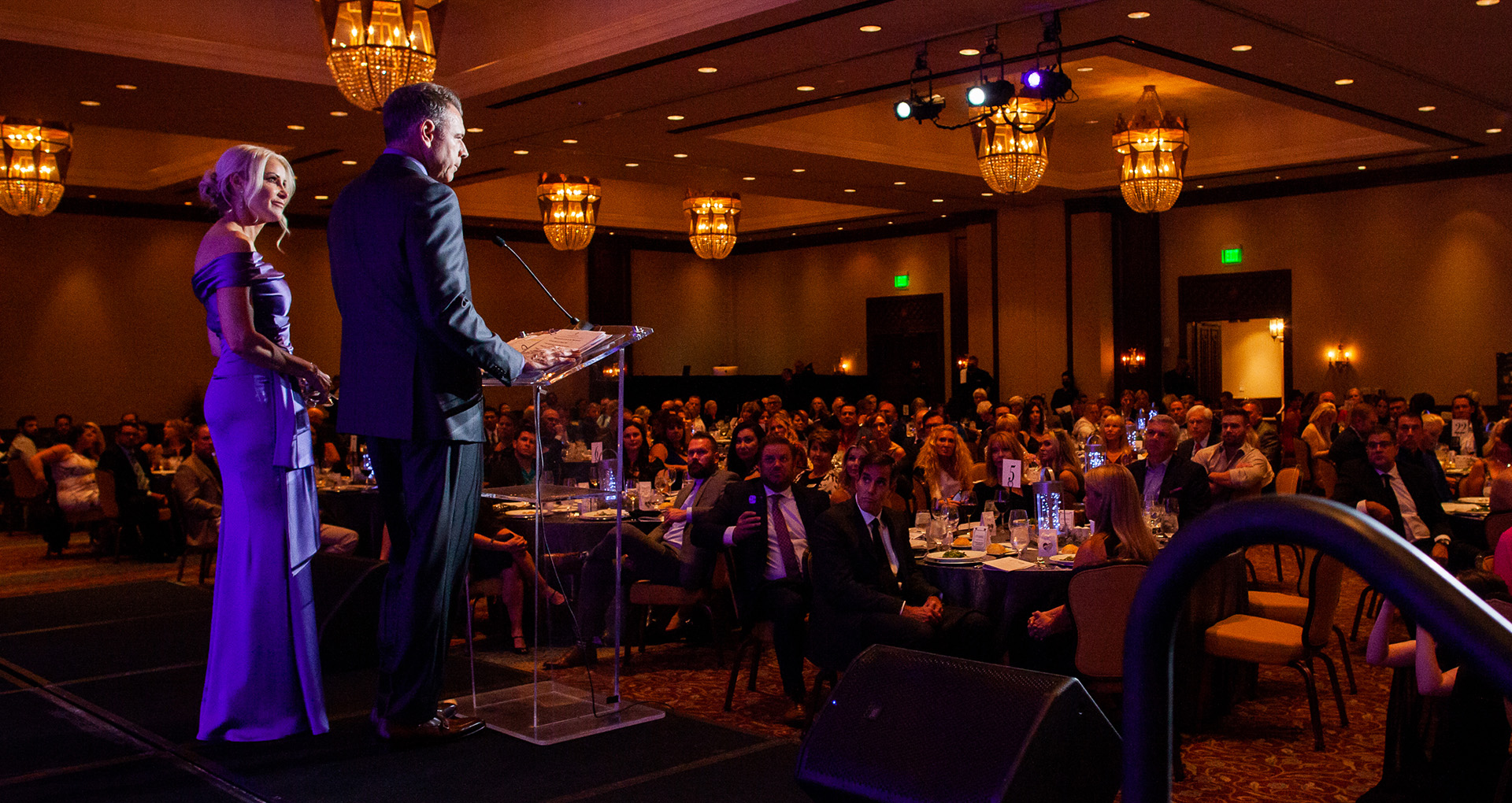 Photo from the 2021 Connections of Hope Gala. 2021 Event Chairs Hugh and Barbara Lytle speak on stage to a ballroom filled with guests.