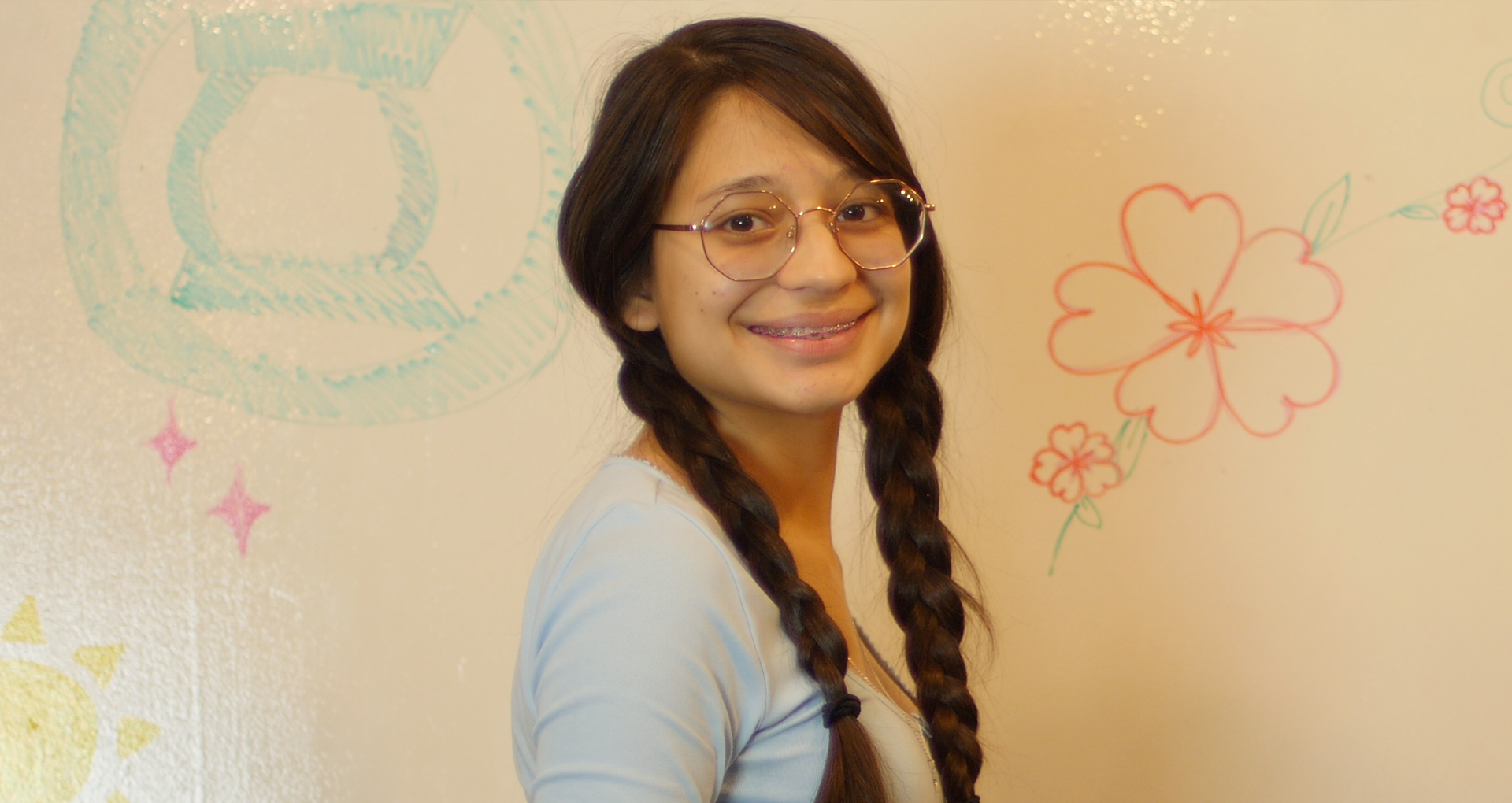 Photo of a teen volunteer smiling at the camera. Behind her are doodles on a whiteboard wall.